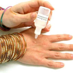 K&ouml;nnte beinhalten: Eine Hand mit mehreren goldenen Armreifen und bunten Armb&auml;ndern wird mit einer klaren Fl&uuml;ssigkeit aus einer kleinen Flasche behandelt. Die Flasche hat Text darauf. Die Fl&uuml;ssigkeit wird auf die Handfl&auml;che aufgetragen.