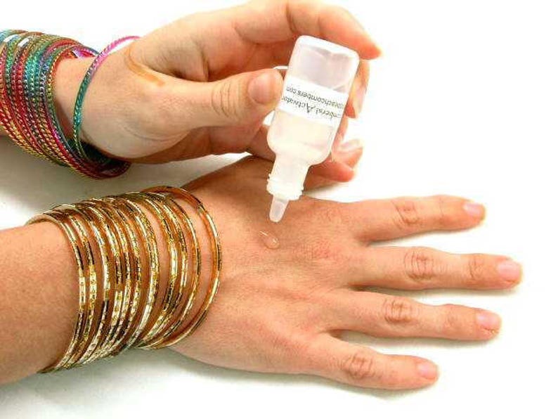 K&ouml;nnte beinhalten: Eine Hand mit mehreren goldenen Armreifen und bunten Armb&auml;ndern wird mit einer klaren Fl&uuml;ssigkeit aus einer kleinen Flasche behandelt. Die Flasche hat Text darauf. Die Fl&uuml;ssigkeit wird auf die Handfl&auml;che aufgetragen.
