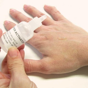 May include: A clear plastic bottle labeled "SUPER ACTIVATOR" is held over a hand. The bottle has a white label with black text. A small amount of liquid is visible on the hand. The background is a plain white surface.