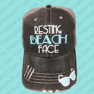 May include: A black and gray trucker hat with a distressed look. The hat has a white mesh back and a white embroidered design that says "Resting Beach Face" with the word "Beach" in light blue. The hat also has a small blue and white sunglasses patch on the side.