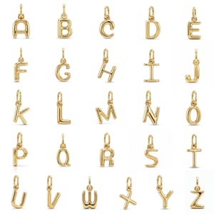 Puede incluir: Una colección de veintiséis dijes de letras individuales de metal en tono dorado, cada uno con una superficie pulida y redondeada y una pequeña argolla en la parte superior para su fijación. El alfabeto se muestra de la A a la Z en cuadrícula.