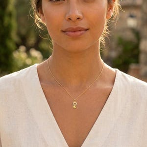 May include: A gold-colored necklace with a small pendant. The pendant has a detailed design and hangs from a delicate chain. The necklace is worn against a white top.