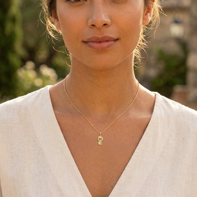 May include: A gold-colored necklace with a small pendant. The pendant has a detailed design and hangs from a delicate chain. The necklace is worn against a white top.