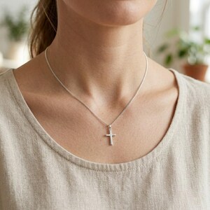 Op de afbeelding: Een zilverkleurige metalen kruishanger hangend aan een fijne bijpassende zilverkleurige ketting. Het kruis heeft een gladde, gepolijste afwerking met een eenvoudig, strak ontwerp en rust op een kledingstuk van neutraal gekleurde stof.