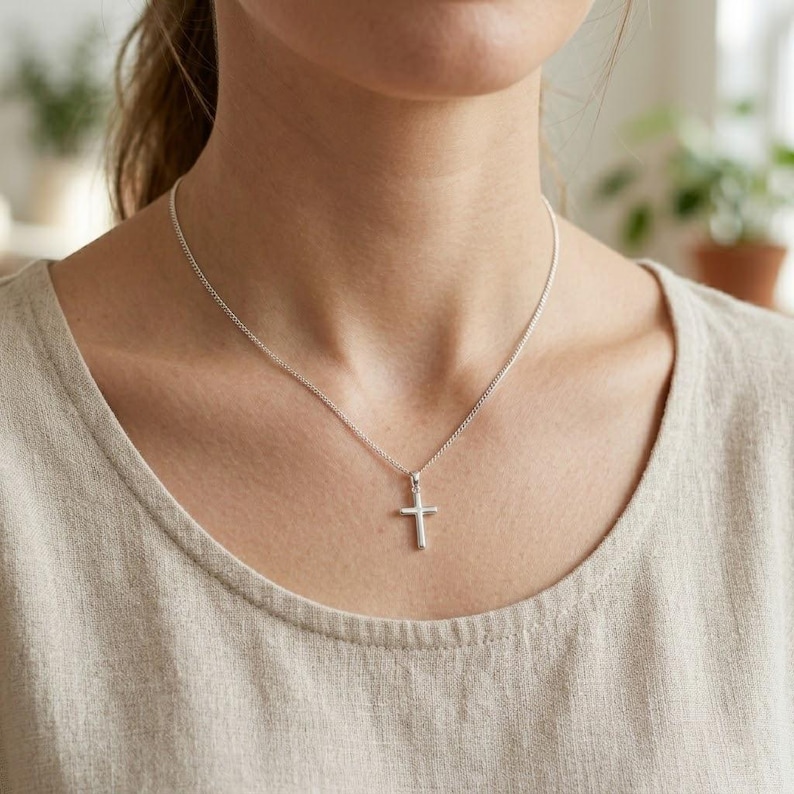 Op de afbeelding: Een zilverkleurige metalen kruishanger hangend aan een fijne bijpassende zilverkleurige ketting. Het kruis heeft een gladde, gepolijste afwerking met een eenvoudig, strak ontwerp en rust op een kledingstuk van neutraal gekleurde stof.