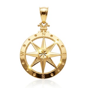 May include: A gold-colored compass pendant with a detailed design. The pendant features a central eight-pointed star, surrounded by a circular frame marked with cardinal directions (N, S, E, W). The pendant has a loop for a chain.