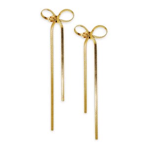 May include: A pair of gold-colored bow earrings with long, dangling chains. The earrings have a bow design at the top and a textured, snake-like chain that hangs down. The earrings are set against a white background.