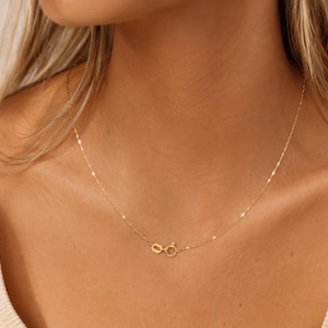 K&ouml;nnte beinhalten: Eine zarte Goldkettenhalskette mit einem d&uuml;nnen, reflektierenden Gliederkettendesign, das am Hals anliegt. Die Halskette wird mit einem kleinen, runden Federringverschluss gesichert, der sichtbar im vorderen Bereich des Schl&uuml;sselbeins positioniert ist.