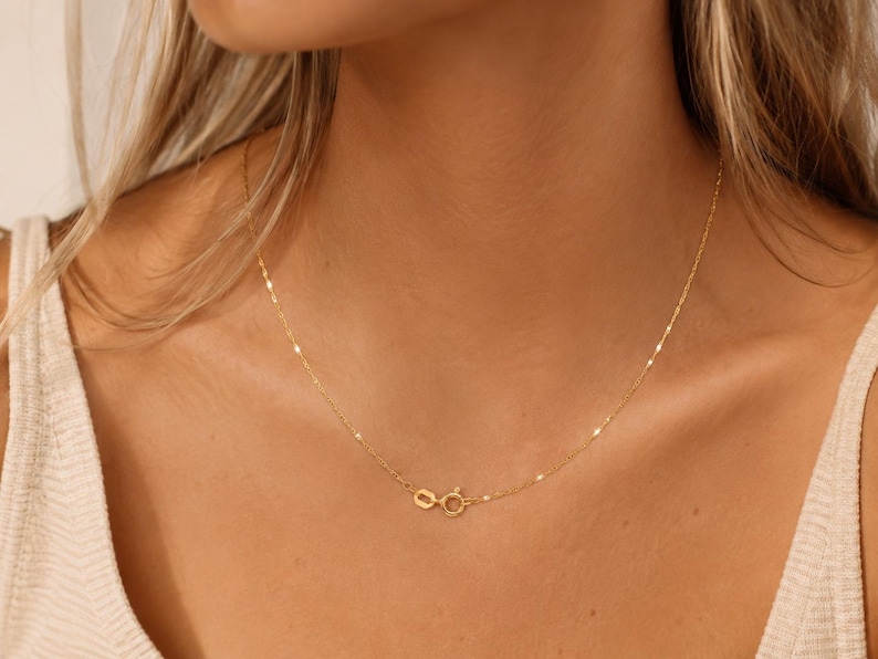 K&ouml;nnte beinhalten: Eine zarte Goldkettenhalskette mit einem d&uuml;nnen, reflektierenden Gliederkettendesign, das am Hals anliegt. Die Halskette wird mit einem kleinen, runden Federringverschluss gesichert, der sichtbar im vorderen Bereich des Schl&uuml;sselbeins positioniert ist.