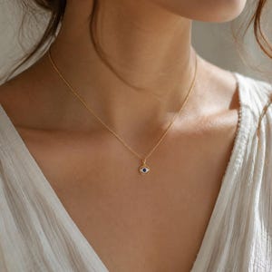 K&ouml;nnte beinhalten: Eine zarte goldene Halskette mit einem kleinen, runden Anh&auml;nger. Der Anh&auml;nger zeigt ein stilisiertes Augenmotiv mit einem dunkelblauen Stein in der Mitte, der von einem wei&szlig;en Emaille-Ring und einem d&uuml;nnen goldfarbenen Metallrand umgeben ist.