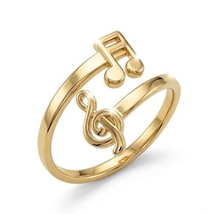 May include: A gold-colored ring featuring musical notes. The ring has a treble clef and a musical note design. The ring wraps around the finger and is made of a shiny, polished metal.