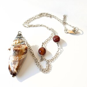 May include: A necklace featuring a brown and white cone-shaped seashell pendant wrapped in silver-toned wire. The silver-toned chain includes two round, polished reddish-brown beads secured with wire wrapping and a lobster claw clasp closure.