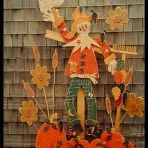 May include: A wooden cutout of a scarecrow with a red shirt, plaid pants, and a straw hat. The scarecrow is holding a sign that says "Keep Out of the Harvest". There are also wooden cutouts of pumpkins, flowers, and cattails.