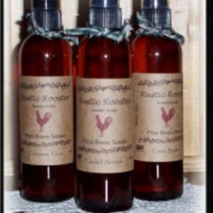 May include: Three glass spray bottles filled with a reddish-brown liquid. Each bottle has a black spray top, a label with a rooster graphic, and a decorative ribbon. The text "Rustic Rooster" is visible on the label.