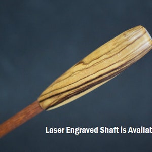 Könnte beinhalten: Nahaufnahme des oberen Teils eines hölzernen Pfeilschafts. Der Schaft besteht aus hellbraunem Holz mit dunkelbraunen Streifen. Der Text "Laser Engraved Shaft is Available" ist am unteren Rand des Bildes sichtbar.