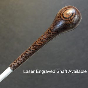 May include: A wooden conductor's baton with a dark brown and black wood grain pattern. The baton has a white shaft and a rounded tip. The text "Laser Engraved Shaft Available" is visible below the baton.