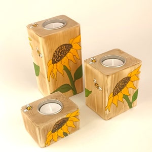 Puede incluir: Tres candelabros de madera con un acabado de vetas de madera natural. Cada soporte presenta un girasol pintado a mano y una peque&ntilde;a abeja. Los candelabros son cuadrados con una hendidura circular para una vela de t&eacute;.