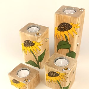 Puede incluir: Juego de cuatro candelabros de madera. Cada soporte es un bloque de madera natural con un dise&ntilde;o pintado de girasol y abeja. Los girasoles son amarillos y marrones, con hojas verdes. Cada soporte tiene un portavelas plateado.