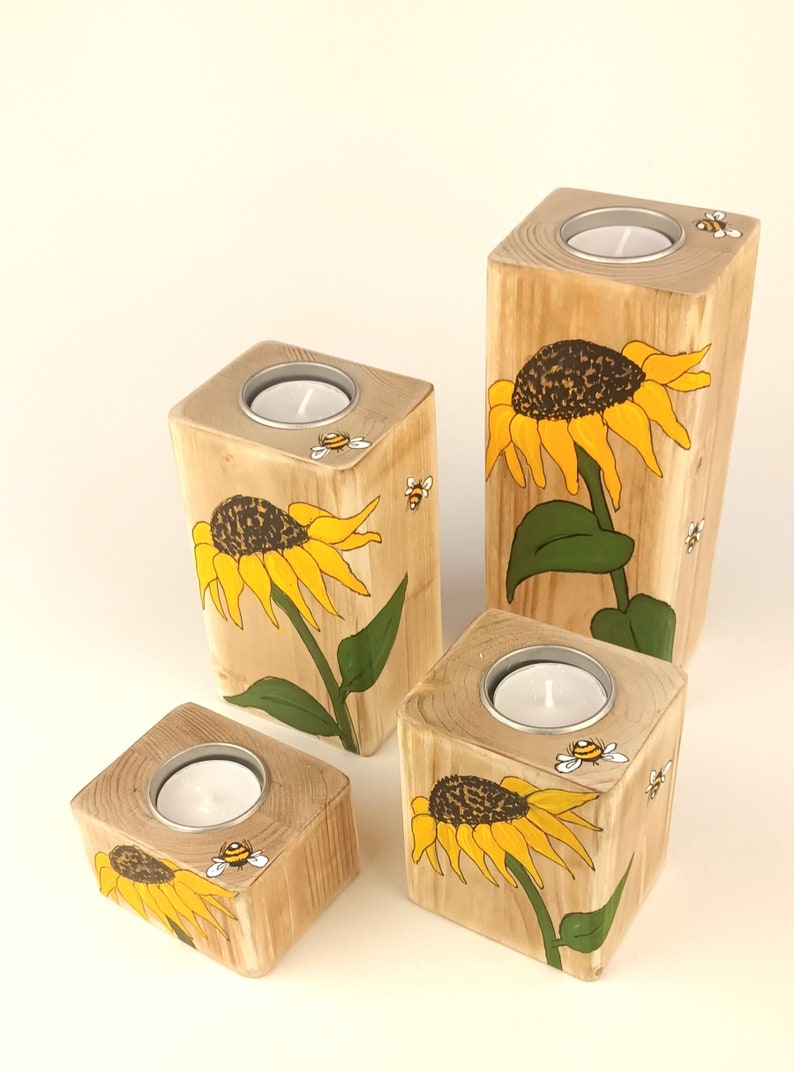 Puede incluir: Juego de cuatro candelabros de madera. Cada soporte es un bloque de madera natural con un dise&ntilde;o pintado de girasol y abeja. Los girasoles son amarillos y marrones, con hojas verdes. Cada soporte tiene un portavelas plateado.