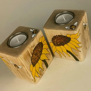 Puede incluir: Dos candelabros de madera con un dise&ntilde;o de girasol y abeja. Cada soporte en forma de cubo tiene una inserci&oacute;n de metal para una vela de t&eacute;. Los girasoles son amarillos y marrones, y las abejas son negras y amarillas.