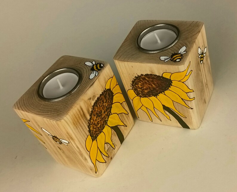 Puede incluir: Dos candelabros de madera con un dise&ntilde;o de girasol y abeja. Cada soporte en forma de cubo tiene una inserci&oacute;n de metal para una vela de t&eacute;. Los girasoles son amarillos y marrones, y las abejas son negras y amarillas.