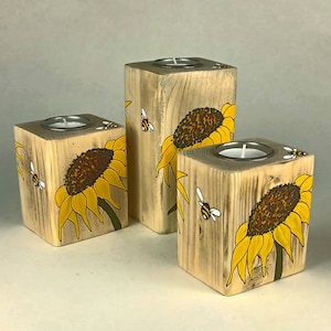 Puede incluir: Juego de tres candelabros de madera. Cada soporte cuadrado tiene una inserci&oacute;n de metal plateado para una vela de t&eacute;. Los bloques est&aacute;n pintados con girasoles amarillos y abejas.