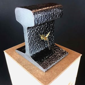May include: A clock constructed from a section of a steel railway track. The metal has a dark, textured, hammered finish on the sides, while the top and bottom edges are smooth and silver. Gold-coloured clock hands are mounted in the centre.