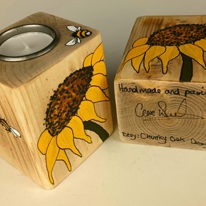 Puede incluir: Dos candelabros de madera con dise&ntilde;os de girasoles y abejas. Uno tiene un inserto met&aacute;lico para una vela de t&eacute;. El otro tiene el texto "Handmade and painted" y una firma. Etsy: Chunky Oak Designs.