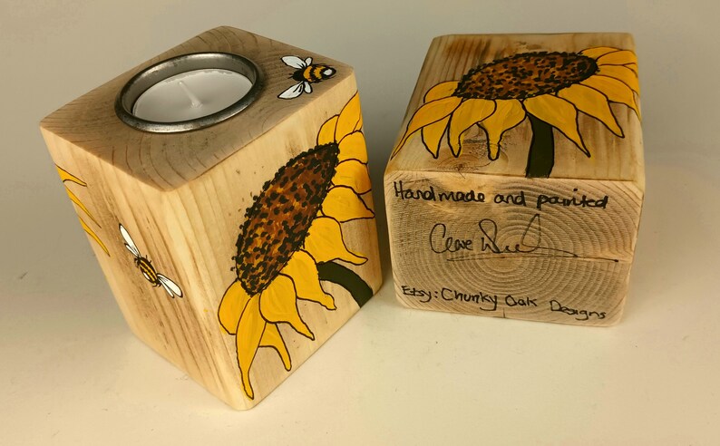Puede incluir: Dos candelabros de madera con dise&ntilde;os de girasoles y abejas. Uno tiene un inserto met&aacute;lico para una vela de t&eacute;. El otro tiene el texto "Handmade and painted" y una firma. Etsy: Chunky Oak Designs.