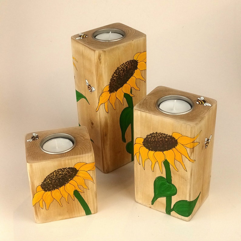 Puede incluir: Juego de tres candelabros de madera. Cada soporte es un bloque de madera natural con un dise&ntilde;o pintado de girasol y abeja. Los girasoles tienen p&eacute;talos amarillos y centros marrones. Cada soporte tiene una inserci&oacute;n de metal para una vela de t&eacute;.