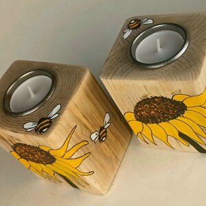 Puede incluir: Dos candelabros de madera con forma cuadrada. Cada soporte presenta un dise&ntilde;o de girasol y abeja, pintado en amarillo, negro y blanco. Los candelabros tienen un inserto de metal para sostener una vela de t&eacute;.