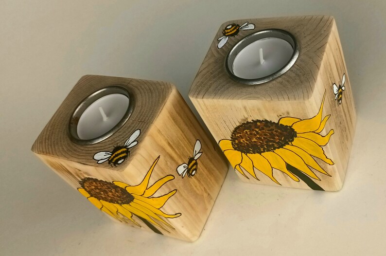 Puede incluir: Dos candelabros de madera con forma cuadrada. Cada soporte presenta un dise&ntilde;o de girasol y abeja, pintado en amarillo, negro y blanco. Los candelabros tienen un inserto de metal para sostener una vela de t&eacute;.
