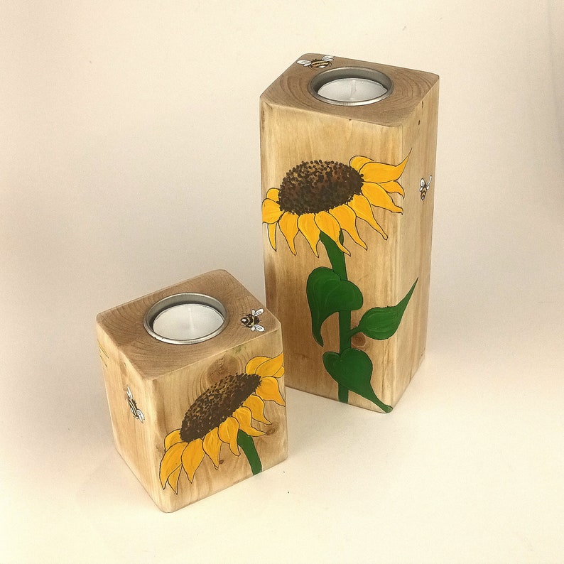 Puede incluir: Dos candelabros de madera, uno rectangular y otro cuadrado. Ambos est&aacute;n pintados con un girasol amarillo y hojas verdes. Peque&ntilde;os portavelas de metal est&aacute;n colocados en la parte superior de cada bloque. Ilustraciones de abejas pintadas en los lados.
