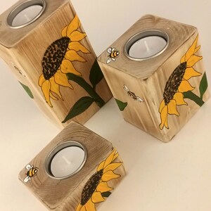 Puede incluir: Tres portavelas de madera con girasoles y abejas pintados a mano. Cada portavelas tiene un inserto met&aacute;lico circular para una vela de t&eacute;. Los girasoles son amarillos y marrones, con hojas verdes. Las abejas son negras y amarillas.
