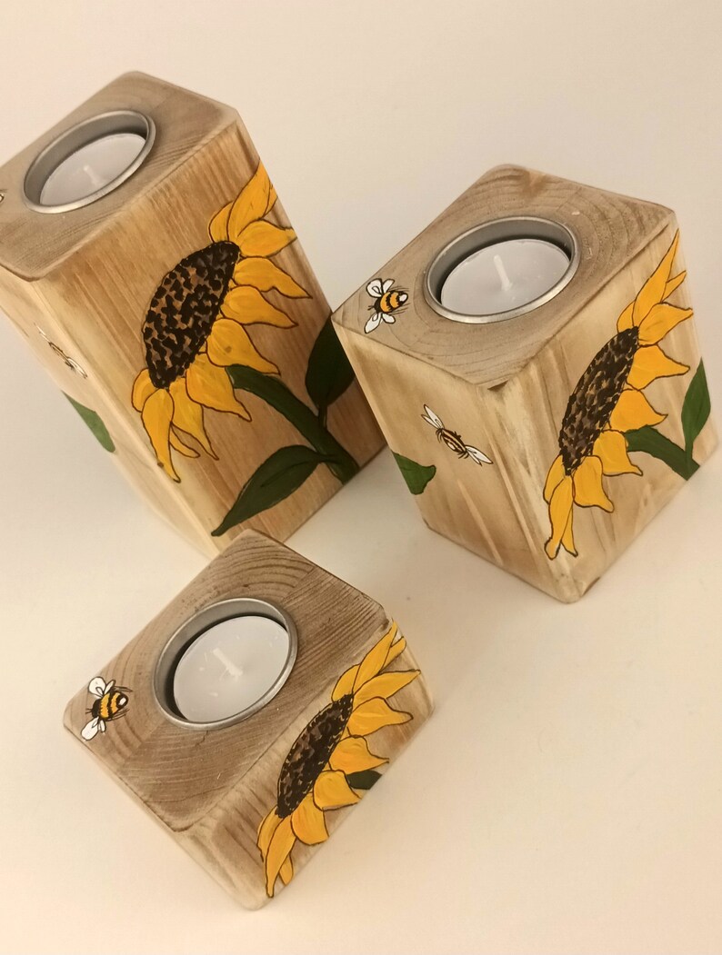 Puede incluir: Tres portavelas de madera con girasoles y abejas pintados a mano. Cada portavelas tiene un inserto met&aacute;lico circular para una vela de t&eacute;. Los girasoles son amarillos y marrones, con hojas verdes. Las abejas son negras y amarillas.