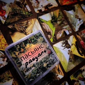 May include: A deck of fortune-telling cards in a clear plastic case. The triangular cards feature vintage illustrations. The case is labelled "Пасьянс pasyans 1910 Fortune Cards". The cards are arranged on a dark wooden surface.