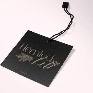 May include: Black square tag with gold lettering that reads "Hemlock Hill" and a small pine tree graphic. The tag is attached to a black string.