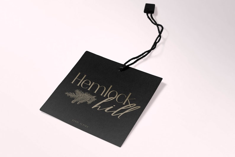 May include: Black square tag with gold lettering that reads "Hemlock Hill" and a small pine tree graphic. The tag is attached to a black string.