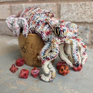 May include: A handmade, multi-colored knitted octopus toy with a button eye, emerging from a small wooden container. Several red and orange dice are scattered around the base, suggesting a tabletop game theme.