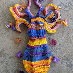 May include: A colorful knitted octopus with a striped body and pink tentacles. The octopus is surrounded by purple and orange dice.