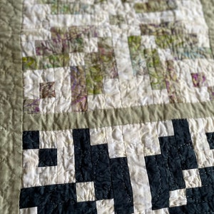 May include: Close-up of a handmade quilt with a patchwork design. The quilt features a combination of white, black, and green fabric squares, creating a geometric pattern. The edges are finished with a sage green border and stitching.