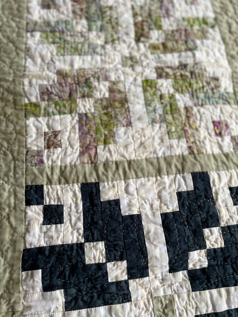May include: Close-up of a handmade quilt with a patchwork design. The quilt features a combination of white, black, and green fabric squares, creating a geometric pattern. The edges are finished with a sage green border and stitching.