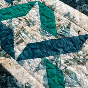 May include: Close-up of a quilted textile with a geometric pattern. The design features teal, dark green, and off-white fabric pieces stitched together. The quilting creates a textured surface with visible stitching lines.