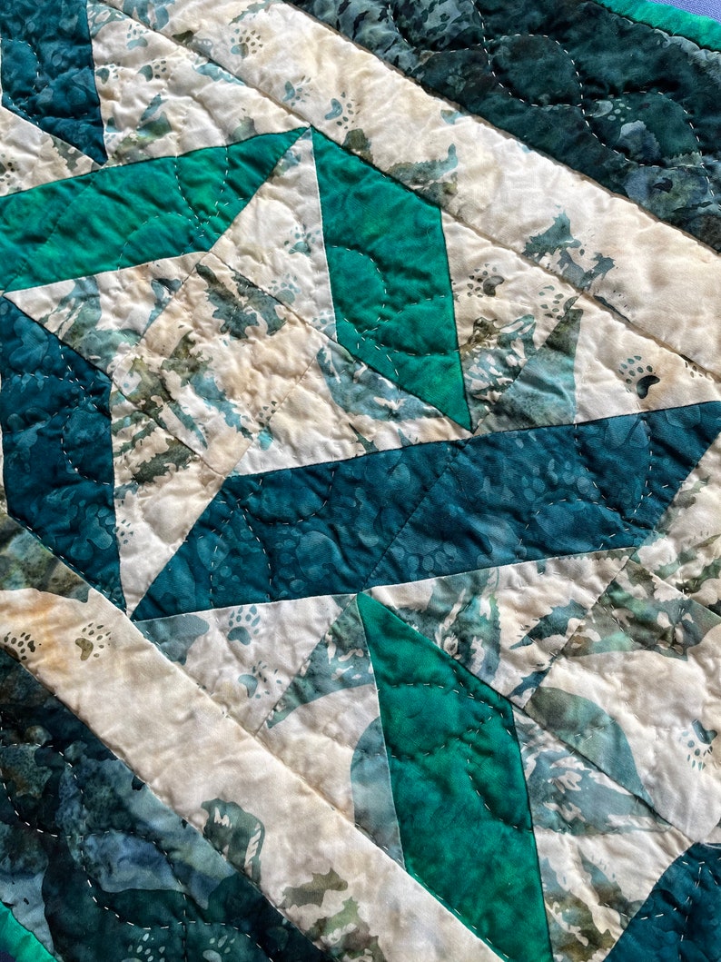 May include: Close-up of a quilted textile with a geometric pattern. The design features teal, dark green, and off-white fabric pieces stitched together. The quilting creates a textured surface with visible stitching lines.