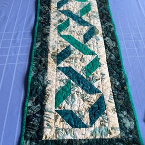 May include: A quilted table runner with a geometric design in shades of teal, green, and cream. The center features a repeating pattern of intersecting shapes, bordered by a darker teal fabric with a scalloped edge and a bright green trim.