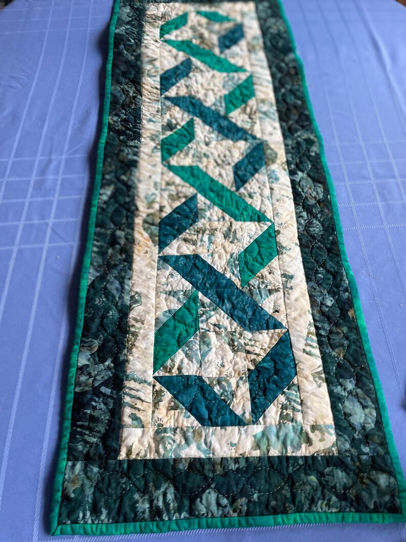 May include: A quilted table runner with a geometric design in shades of teal, green, and cream. The center features a repeating pattern of intersecting shapes, bordered by a darker teal fabric with a scalloped edge and a bright green trim.