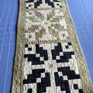 May include: A rectangular quilted table runner with a repeating pattern of black and white geometric shapes. The centre section features a floral print in shades of green, purple, and white. The border is a muted green.