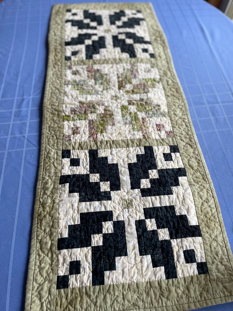 May include: A rectangular quilted table runner with a repeating pattern of black and white geometric shapes. The centre section features a floral print in shades of green, purple, and white. The border is a muted green.