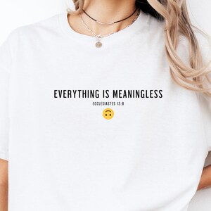 Può includere: Maglietta bianca con il testo "EVERYTHING IS MEANINGLESS" e "ECCLESIASTES 12:8" stampato sul davanti. Un'emoticon gialla capovolta si trova sotto il testo.