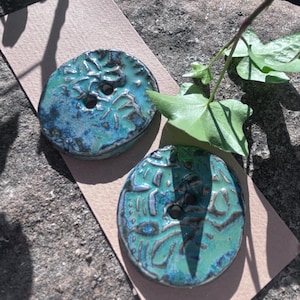 May include: Two round ceramic buttons featuring a textured, glossy glaze in shades of turquoise, teal, and deep blue. Each button has a single central hole and an irregular, organic surface pattern with dark, recessed lines throughout.